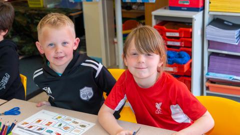 Boy and a girl sat at their desk with worksheets smiling at the camera